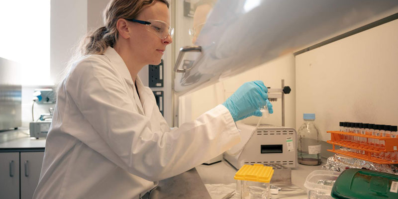 A female researcher works at a microscope in the BioArCh labs, she's wearing a white labcoat and blue latex gloves.