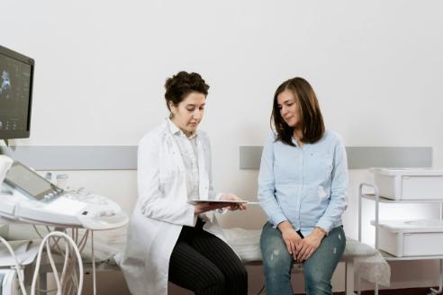 A female clinician with brown hair and white skin looks at a tablet with a female patient with brown hair wearing a light blue blouse and blue jeans They are sat in a consulting room.