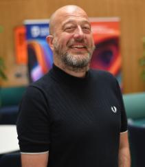 A man with white skin, bald head, brown eyes, dark grey moustache and beard wearing a black t-shirt and smiling to the camera.