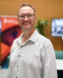 A man with white skin, light brown hair, a grey beard, black glasses wearing a white and brown printed shirt smiling to the camera.