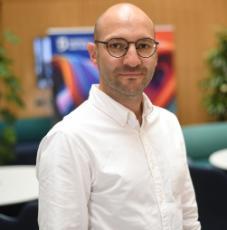 A man with tanned skin, brown hair and bald head wearing black glasses and a white shirt smiling to the camera