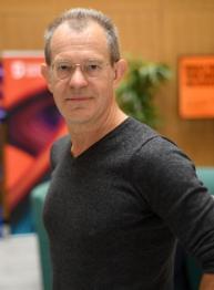 A man with white skin, grey hair and black glasses wearing a charcoal v-neck woollen jumper looking to the camera.