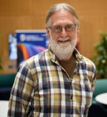 A man with white skin, light blond hair, blue eyes, white beard and moustache and glasses wearing a light brown t-shirt and black, brown and white checked shirt smiling to the camera.