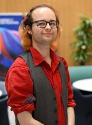 A man with white skin, blue eyes, brown shoulder length hair, a black stud in one ear, black glasses wearing a red shirt and a dark grey waistcoat smiling to the camera.