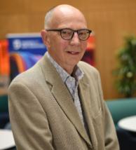 A man with white skin, grey hair and black glasses wearing a camel-coloured jacket and a blue shirt smiling to the camera.