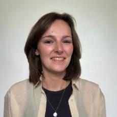 A woman with white skin, brown eyes and hair wearing a black vest underneath a cream blouse smiling directly at the camera.