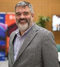 A man with white skin, grey hair and grey moustache and beard wearing a grey and white houndstooth jacket over a light blue shirt smiling to the camera.