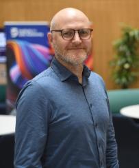 A man with white skin, bald head, grey moustache and beard wearing a blue shirt smiling to the camera.