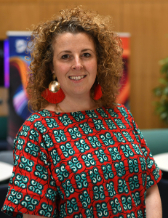 A lady with white skin, brown curly shoulder length hair with brown eyes wearing a blue and red patterned short sleeve dress and gold metal and red tassel earrings.