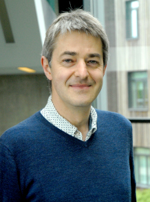 A man with white skin, grey hair wearing a blue v-neck jumper over a blue shirt smiling to the camera.