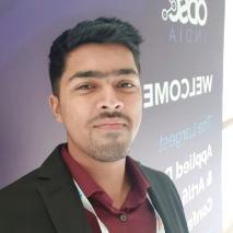 A man with brown skin, black hair and brown eyes wearing a maroon shirt and black jacket smiling to the camera.