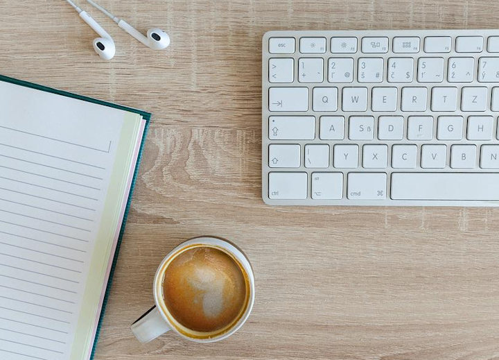 A computer keyboard, notebook, ear pods and a cup of coffee on a table