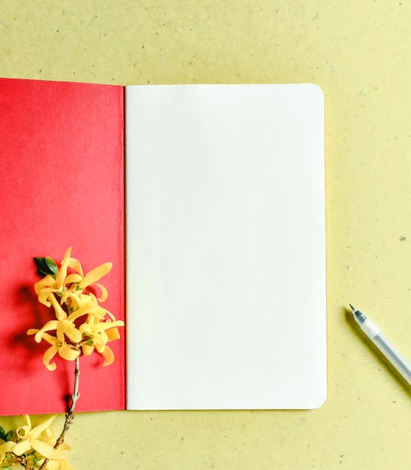 An open unlined notebook with a red cover, a pen next to it and a yellow flower