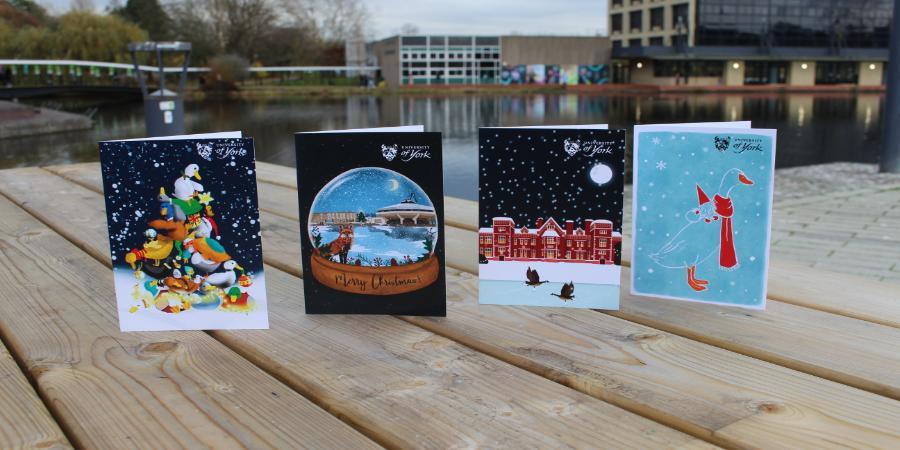 The image features Christmas card competition winners from previous years on Greg's Place bench with the campus lake and Exhibition Centre building featured in the background