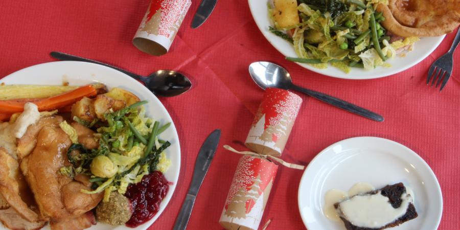 The image features a vibrant spread of Christmas lunches, alongside a desert plate and Christmas crackers placed on the red coated table with cutlery.