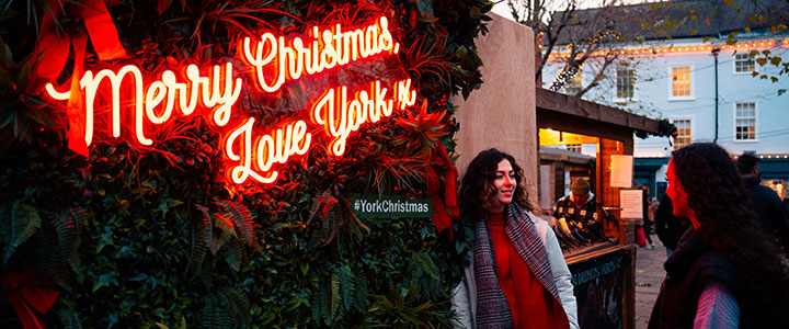 Two students at the York Christmas market, standing in front of an illuminated sign reading 'Merry Christmas Love York'