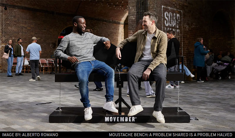 Two men talking on a Movember-branded bench. Text reads: Image by Alberto Romano. Moustache bench: a problem shared is a problem halved.